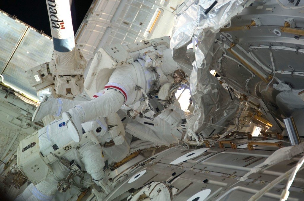Two astronauts in white spacesuits on a spacewalk outside the International Space Station.