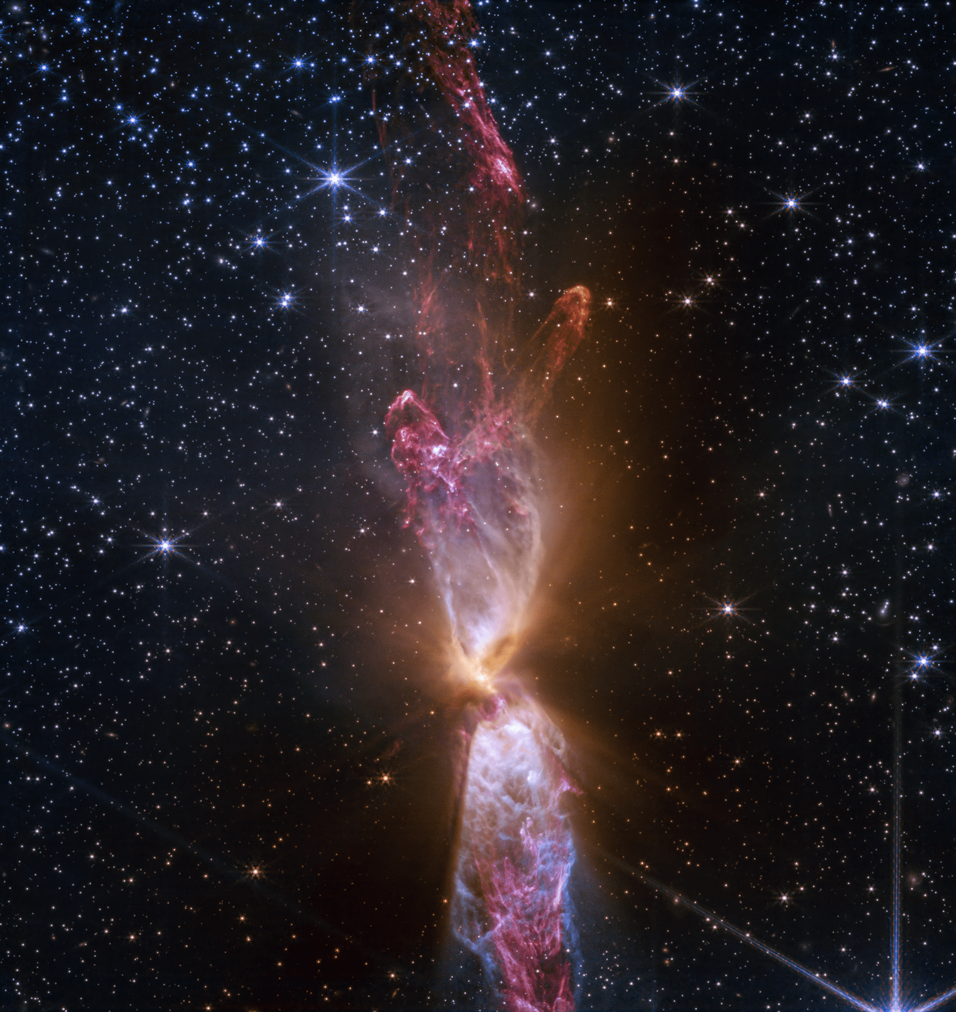 At the center is a thin vertical cloud known as Lynds 483 that is shaped like an hourglass with irregular edges. At lower center are two discrete bright white, tiny blobs of light that have raced away from the hidden central stars. The top lobe shows a more prominent orange U-shape. Orange bleeds into light purple, and brighter pink at its edges. Some background stars are visible through sections of this lobe. Higher up, there is an orange arc. Some brighter pink material extends to the top edges near the center. In the lower lobe, less orange is visible. More opaque light purple is in its top third, rippling out into semi-transparent blues and pinks. The lower lobe has more texture. V-shapes left and right of the lobes are darkest, and the background stars in these areas appear orange. Elsewhere, the black background of space is clearer, speckled with tiny white stars and faint orange galaxies.