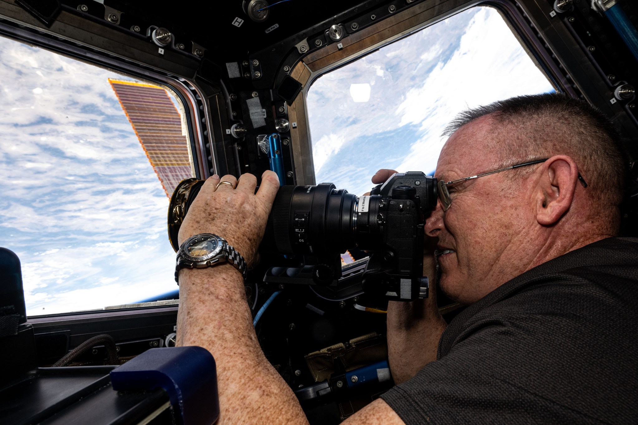 Wilmore, wearing a black polo shirt and a watch on his left wrist, holds a camera to his face with his right hand and adjusts the lens with his left. He is facing one of the cupola windows and a second window is visible to his right. A cloudy Earth below and a bit of one of the station&rsquo;s solar panels can be seen through the windows.