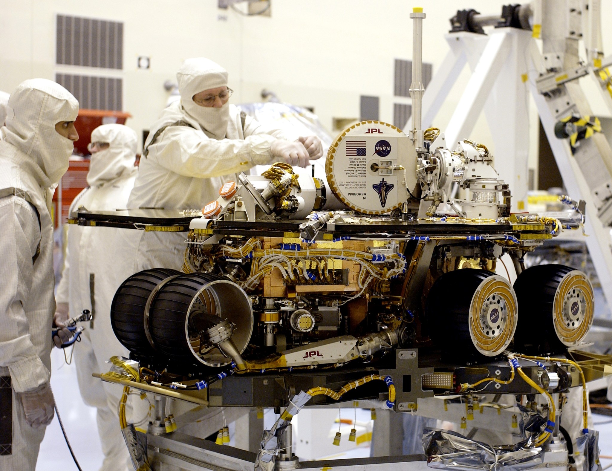 Technicians in white jumpsuits that cover everything but their eyes and nose work on the Spirit rover. The rover has four visible wheels, covered by a rectangular platform. There are many yellow and white cords all over the rover. There is a circular plaque attached to a white antenna.