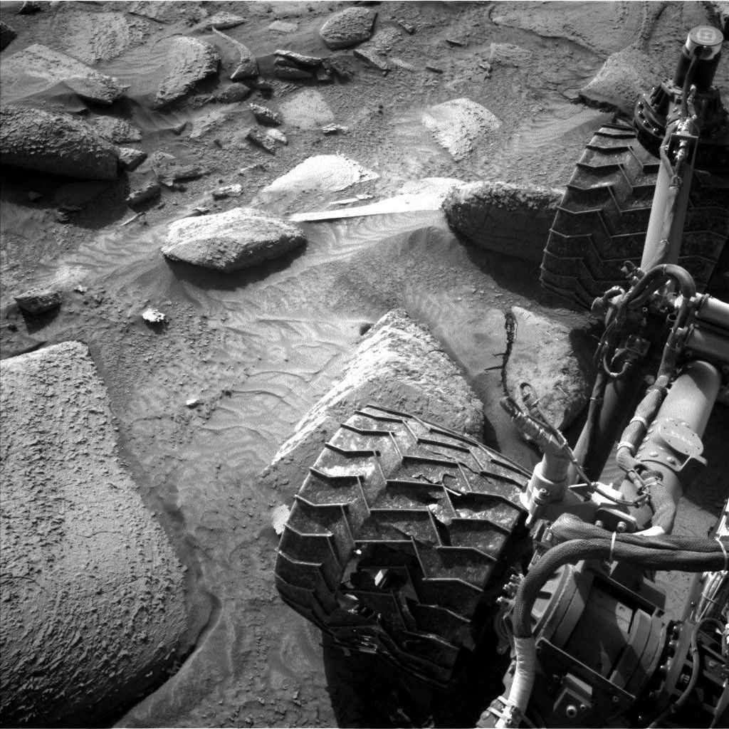 A grayscale photo from the Martian surface shows very uneven terrain covered in large rocks, with the Curiosity rover taking up the right side of the image, from the top-right corner to the bottom center of the frame. Parts of the rover chassis are visible, as well as two of the wheels; the one near the top of the frame is seen perched atop one of the large rocks here.
