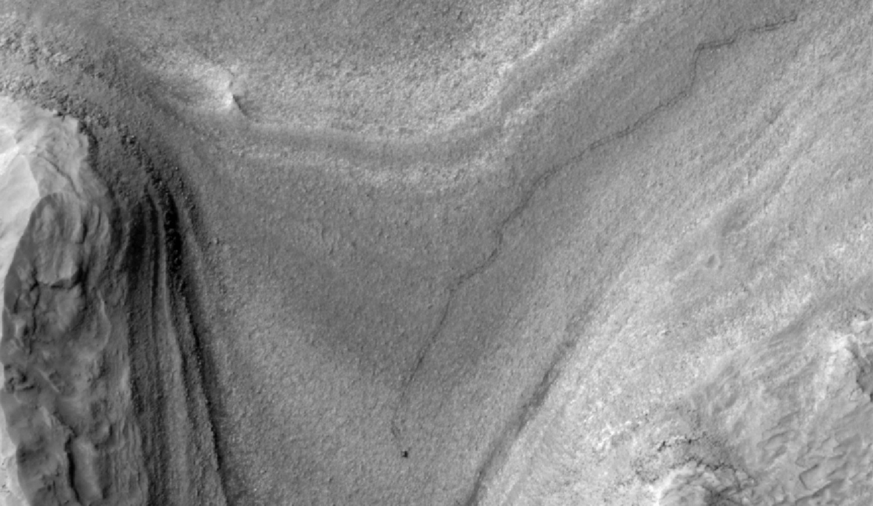 High-resolution grayscale image of Mars showing layered terrain with smooth slopes, rocky ridges, and a small dark speck near the center, identified as NASA's Curiosity rover