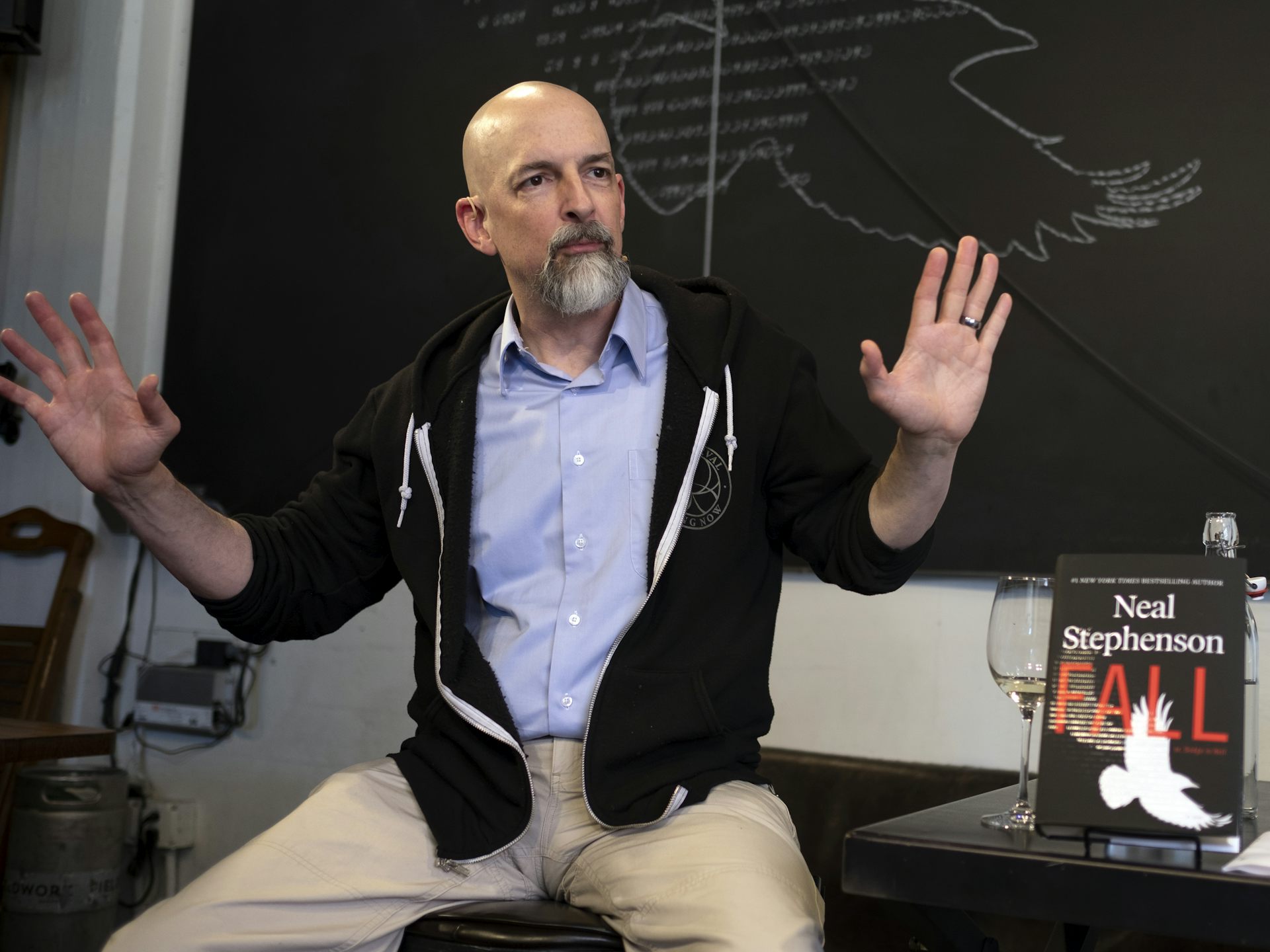 Photo of bald man with goatee sitting on a desk with both hands raised.