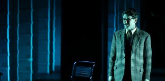 Eliza, a theatrical thriller about the first chatbot, feels emotionally distant. That’s the point A man in a suit stands on a darkened stage with a grid of stacked document boxes behind him.